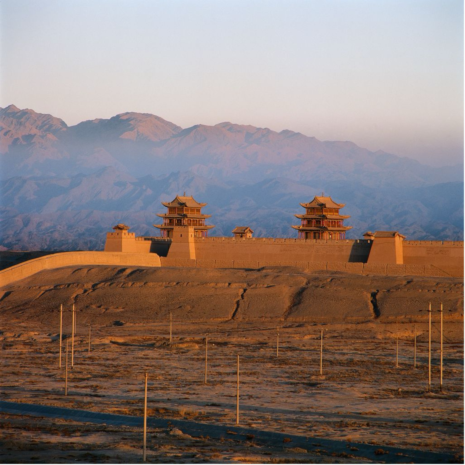 Festung Jiayuguan mit zwei Tortürmen im Abendlicht, dahinter das Qilian-Gebirge, im Vordergrund Wüstenebene mit Strommasten