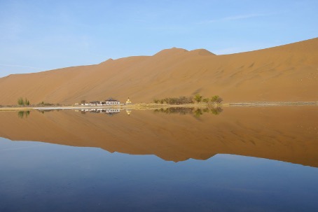 Hohe Sanddünen der Badain-Jaran-Wüste spiegeln sich in einem stillen See, am Ufer ein kleiner Tempel