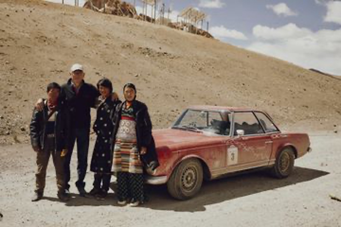 Rallye-Teilnehmer mit Einheimischen in traditioneller tibetischer Kleidung neben einem roten Mercedes-Oldtimer mit Startnummer 3 im Hochland